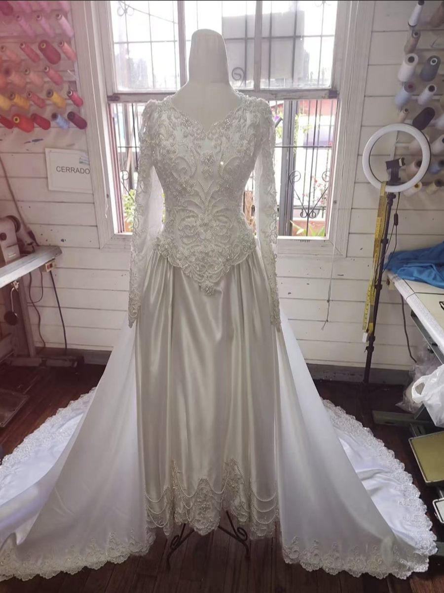 Vestido de novia en satén marfil con corpiño bordado en perlas, mangas de encaje y cola larga, montado en maniquí dentro del taller