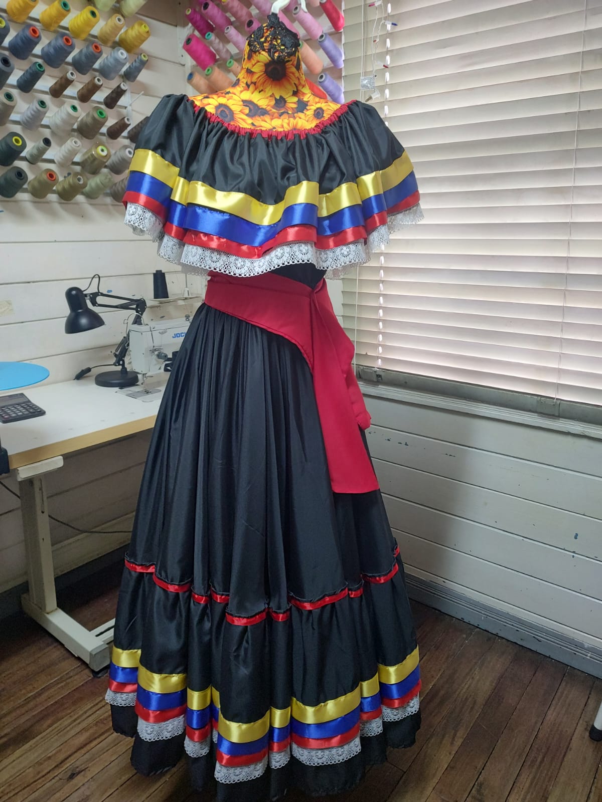 Traje típico en tafetán negro con escote de flores amarillas, holán de cintas rojas, amarillas y azules bordeado en encaje, falda de vuelos con las mismas cintas y faja roja al frente, montado en maniquí dentro del taller